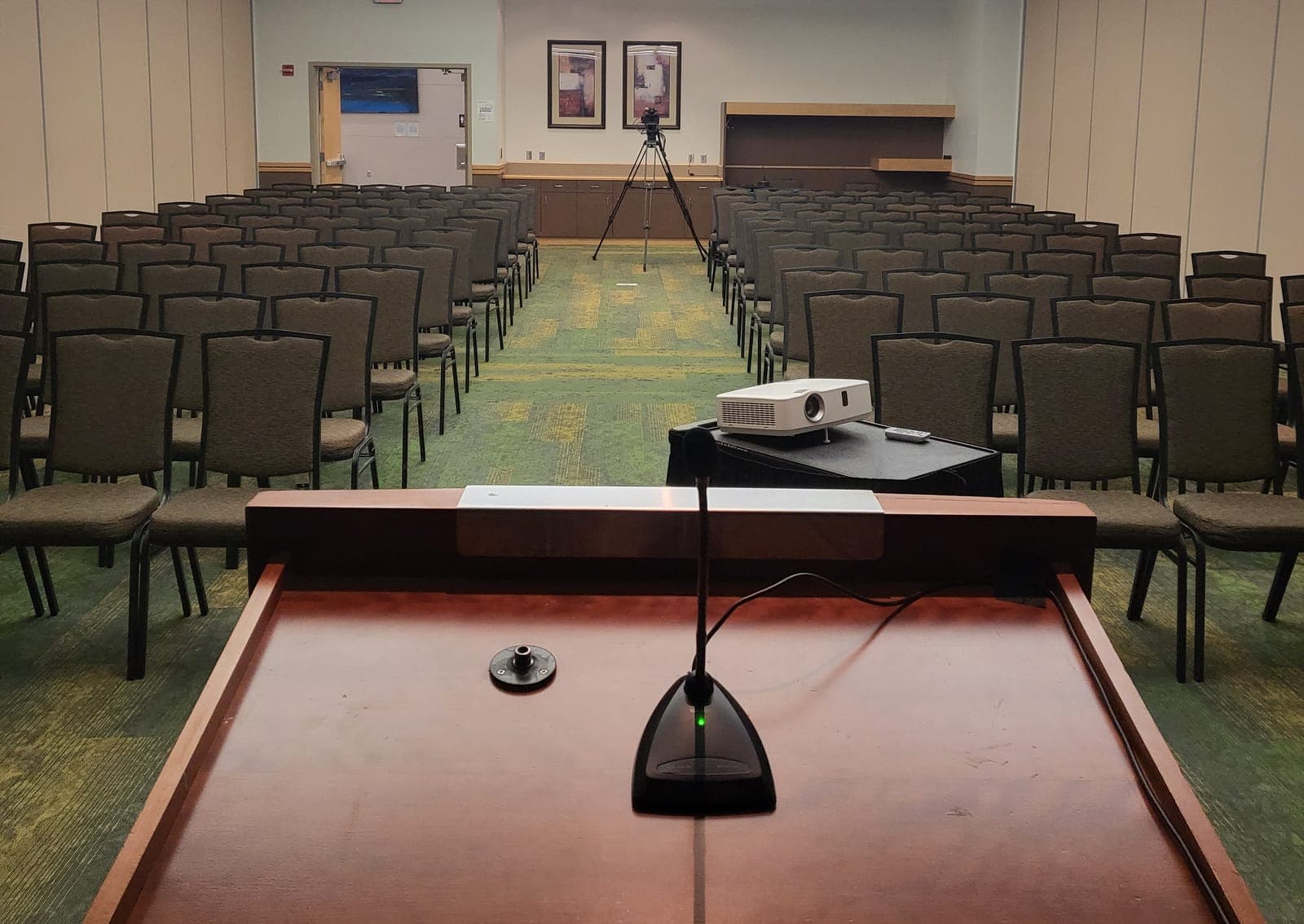 The view from the lectern in an empty session room: a microphone in the foreground, rows of chairs, a projector, and a video camera on a tripod